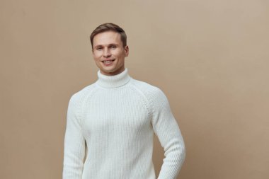 Kısa saçlı, rahat, beyaz balıkçı yaka kazak giyen neşeli genç bir adamın portresi basit bir bej arka planda duruyor. Görüntü sıcak ve dostane bir atmosfer yakalıyor.