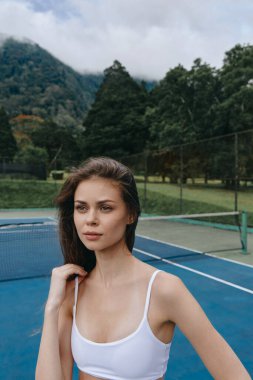 Etrafı yeşilliklerle çevrili bir tenis kortunda poz veren kendine güvenen genç kadın atletizmi ve açık hava sporlarının ruhunu yakalıyor.