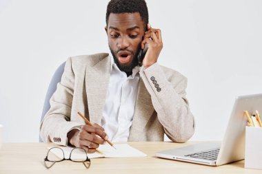 Kendine güvenen, bej ceket giyen ve iş yerinde telefonla konuşan beyaz gömlekli genç bir adam. Şaşırmış ya da heyecanlı görünüyor, dizüstü bilgisayar ve defterle temiz bir çalışma alanına oturmuş,