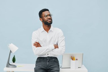 Sakallı, gözlüklü, resmi beyaz gömlekli, kolları masanın yanında diz üstü bilgisayar, sanat malzemeleri ve açık mavi arka planda lambayla çapraz duran kendine güvenen bir adam. Konsept: Modern ofis ortamı