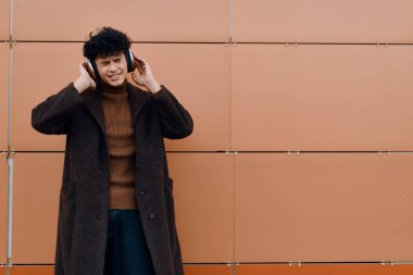 Turuncu duvarın önünde kulaklıkla müzik dinleyen Asyalı bir adam. Müzikten hoşlanan genç bir adam. Moda şehir yaşam tarzı konsepti.