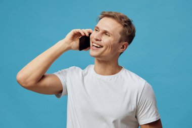 Gülümseyen, beyaz tişörtlü, cep telefonuyla konuşan genç beyaz bir adam mavi arka plan dijital göçebe yaşam tarzı konsept iletişim teknolojisi kablosuz bağlantısına karşı duruyor.