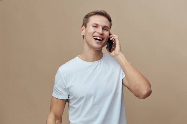 Kısa saçlı neşeli genç adam beyaz bir tişört giyiyor, gülümsüyor ve cep telefonuyla konuşuyor, bej bir arka plana karşı duruyor, samimi bir iletişim anını yakalıyor ve...