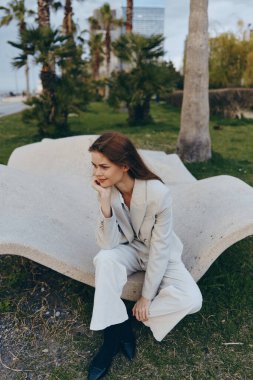 Açık bej renkli takım elbiseli düşünceli bir kadın modern bankta oturuyor. Palmiye ağaçları ve yeşilliklerle çevrili. Sonbahar sezonu.