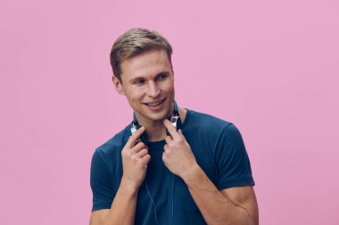 Kulaklıklı gülümseyen adamın portresi, günlük mavi tişört, neşeli ifade, genç yetişkin, pembe arka planda izole edilmiş, stüdyo çekimi, rahatlatılmış ruh hali, yaşam tarzı konsepti, pozitif titreşim, modern