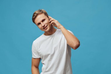 Beyaz tişört giyen gülümseyen beyaz bir adam akıllı telefonda dijital göçebe yaşam tarzını ve uzaktan çalışma kavramını gösteren modern iletişim teknolojisini anlatırken mavi arka planda duruyor.