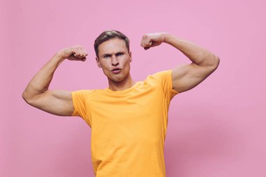 Kaslı, sarı tişört giymiş kaslı bir gencin portresi. Pembe bir arka plana karşı durarak güç ve zindelik göstermek için pazılarını esnetiyor. Sağlık ve spor salonu için ideal.