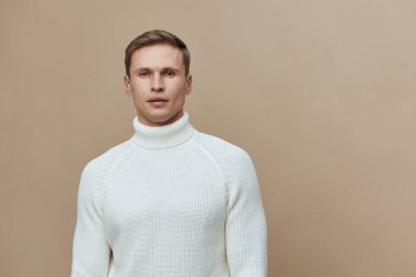 Beyaz balıkçı yaka kazak giyen ciddi bir adam bej arka planda, tarafsız bir yüz ifadesine karşı duruyor. Günlük kışlık giysiler giyiyor, sakin ve özgüvenli. Kavram: minimalist