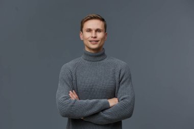 Gri balıkçı yaka kazak giymiş mutlu genç adam stüdyo çekimlerinde gri arka planda gülümsüyor. Kendine güveni tam bir erkek model, rahat ve misafirperver tavırları var. Yakın plan portre
