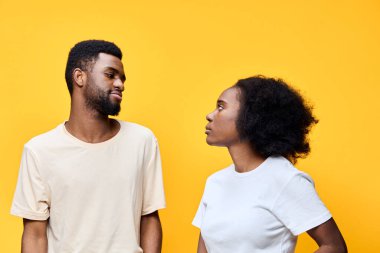 Neşeyi ve bağlantıyı temsil eden, parlak sarı bir arka planda karşılıklı bakışlar atan genç bir çift, güncel moda eğilimlerini yansıtan gelişigüzel giyinmişlerdir.