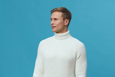 Açık tenli, beyaz örgülü balıkçı yaka kazak giyen, yavaşça gülümseyen ve arkasında düz mavi bir arka planı olan genç bir adamın yakın çekim stüdyo portresi. Konsept: gelişigüzel