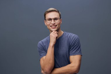 Mavi tişört giymiş gözlüklü mutlu bir genç adam gülümsüyor ve gri arka planda kameraya bakıyor. Konsept: Güven ve pozitiflik.