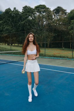 Beyaz etekli ve spor sutyenli enerjik bayan tenisçi, yemyeşil yemyeşil ile çevrili güneşli bir kortta güven ve neşe saçıyor.