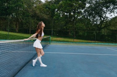 Beyaz spor kıyafetleri içinde kendine güvenen genç bir bayan tenisçi, yemyeşil bir zemine karşı duruyor, neşe ve aktif bir yaşam tarzı sunuyor, spor promosyonu için ideal.