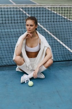 Şık spor kıyafetleri içinde tenis kortunda oturan genç bir atletik kadın özgüven ve kararlılık sergiliyor, fitness temalı projeler için mükemmel.