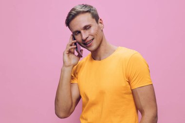 Kısa saçlı, sarı günlük tişörtlü neşeli bir genç adam. Pembe arka planda, kapalı bir ortamda akıllı telefondan mutlu bir şekilde konuşuyor. Neşe ve iletişim, yaşam tarzı kavramı aktarıyor..