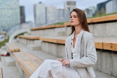 Bej renkli hafif takım elbiseli iş kadını dışarıda beton basamaklarda bulanık modern şehir arka planına karşı oturuyor profesyonel bir atmosferde düşünceli ve ciddi duygular ifade ediyor..