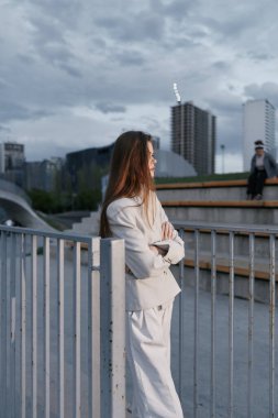 İş elbiseli kadın şehir binalarına ve bulutlu gökyüzüne karşı düşünceli bir şekilde dışarıda duruyor, yansıma, profesyonellik ve sakin bir ruh hali sergiliyor..