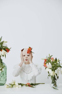 Genç bir kadın, başı ellerinde, çiçeklerle süslenmiş, temiz, beyaz bir arka planda çiçek aranjmanlarıyla çevrili narin beyaz bir elbise giyiyor.