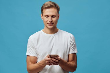 Kısa saçlı neşeli genç adam düz beyaz bir tişört giyiyor, cep telefonuyla meşgul, aşağıya bakarken mutlu bir şekilde gülümsüyor, düz mavi stüdyo arkaplanı, rahat ve rahat bir ruh hali, teknoloji
