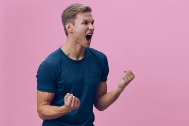 Enerjik genç bir adam, günlük mavi tişörtleriyle, yüz ifadesi ve vücut diliyle coşku ve neşe gösteriyor, canlı pembe bir arka plan karşısında duruyor, stüdyo çekimi, mutluluğu taşıyor.