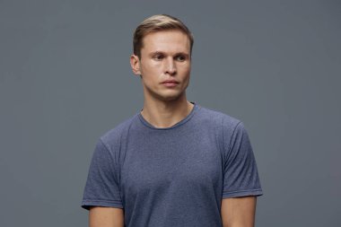 Gri arka planda izole edilmiş ciddi bir ifadeye sahip, günlük mavi tişörtlü, düşünceli beyaz bir adam. Duygu, yalnızlık, zihinsel sağlık ve dijital detoks kavramı