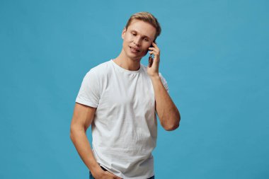 Beyaz tişörtlü genç adam mavi arka planda el ele dururken akıllı telefonla konuşuyor. Modern günlük tarz dijital göçebe iletişim için cep telefonunu uzaktan kullanıyor