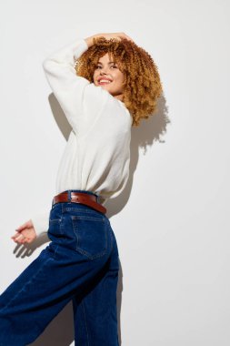 Şık ve kendine güvenen kıvırcık saçlı, beyaz bir kazak ve kot pantolon giyen, temiz, açık bir arkaplana karşı poz veren modern moda eğilimlerinin ve bireysel eğilimlerinin mükemmel bir temsilcisi olan bir kadın.