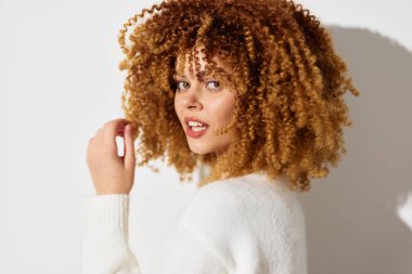 Kıvırcık saçlı, neşeli yüzlü bir kadın, şık bir şekilde poz veriyor, doğal güzelliği ve kendine güveni vurguluyor, yumuşak, hafif bir arka plana karşı canlı kıvrımlar sergiliyor,