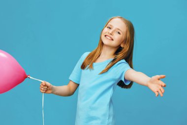 Açık mavi tişörtlü, uzun kızıl saçlı neşeli bir kız pembe bir balonla parlak mavi bir arkaplana poz veriyor, neşeli çocukluk anılarının özünü yakalıyor.