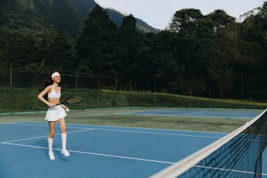 Beyaz spor kıyafetleri içinde neşeli bayan tenisçi mavi tenis kortunda duruyor, etrafa şevk ve zarafet saçıyor yeşil ağaçların arasına, spor reklamcılığı için mükemmel.