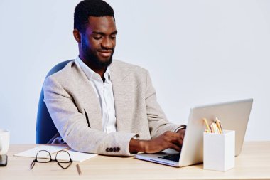Kendine güvenen Afro-Amerikan bir adam, parlak çağdaş bir ofiste dizüstü bilgisayar kullanıyor. Dijital teknolojiyle uğraşırken odaklanma ve profesyonellik sergiliyor.