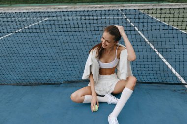 Şık beyaz spor kıyafetli genç bir atletik kadın tenis kortunda rahatlıyor, tenis topu tutarken kendine güven ve neşe saçıyor.