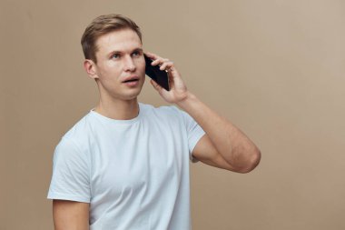 Cep telefonundan konuşan beyaz bir adam bej arka planda izole edilmiş beyaz tişört giyiyor. Mobil teknoloji ve iletişim araçlarıyla uzaktan çalışan dijital göçebe yaşam tarzına odaklanıyor.