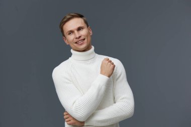 Kısa saçlı, neşeli bir gülüşü olan, beyaz balıkçı yaka kazak giyen, gri arka planda güvenle duran, rahatlığı, stili ve pozitif enerjiyi ön plana çıkaran erkek model. Kavram