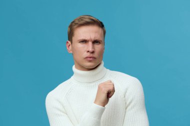 Beyaz balıkçı yaka kazaklı kafası karışmış genç bir adam mavi zemine karşı şüpheli bir yüz ifadesiyle poz veriyor. Düşünüyor ya da sorguluyor, gündelik tarzda. Kavram: belirsizlik veya şüphe
