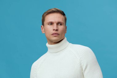 Yakından bakınca, beyaz balıkçı yaka kazak giyen yakışıklı bir adam ciddi bir ifadeye sahip, sağlam mavi bir arka plana bakıyor. Moda, tarz, erkek kıyafeti, kışlık kostüm için mükemmel.