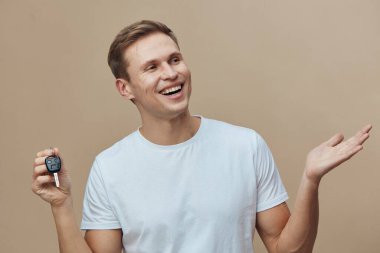 Kısa kahverengi saçlı, günlük beyaz tişörtlü, uzaktan kumandayla hareket eden ve gülümseyen, bej arka planda duran neşeli genç adam. Mutlu anlamlı poz, yaşam tarzı