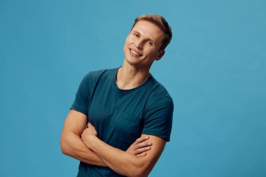 Mavi tişörtlü, kollarını çapraz bağlamış gülümseyen beyaz bir adam kameraya bakıyor. Mavi arka plandaki pozitif duyguları gösteriyor. Günlük tarz ve dijital detoks teması.