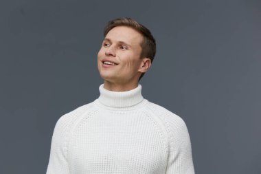 Kısa kahverengi saçlı, rahat beyaz balıkçı yaka kazak giyen, gülümseyen ve gri arka planlı profesyonel bir stüdyo ortamında yüzünü çeviren mutlu genç adam. Kavram: pozitiflik, gelişigüzel