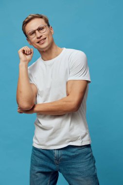 Kısa saçlı, gözlüklü, sıradan kıyafetli, gülen ve arkadaş canlısı tavırlarıyla poz veren mutlu bir genç. Sade mavi arka planda izole edilmiş, rahat ve pozitif ruh hali, günlük giysiler. Kavram: Gençlik