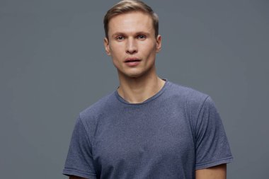 Kısa saçlı, açık tenli, mavi tişörtlü, tarafsız gri bir arkaplanda duran genç bir adamın portresi. Stüdyo çekimi doğal yüz ifadeleri, günlük tarz, minimalist