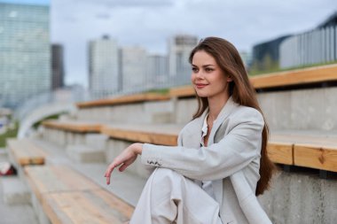 Açık renk takım elbiseli kadın şehir manzarası arka planında şehir ahşap merdivenlerinde oturuyor, gülümsüyor ve rahatlıyor, modern moda ve iş yaşamını somutlaştırıyor.