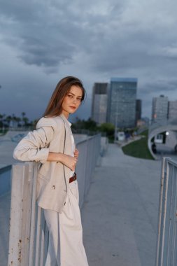 Bej takım elbiseli, şehir binaları ve arka planda bulutlu gökyüzü ile metal parmaklıkların yanında poz veren kendine güvenen genç bir kadın iş profesyonelliği ve sakin duygular sergiliyor..