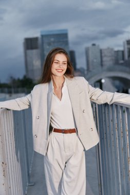 Açık bej renkli takım elbiseli, kendine güvenen bir kadın, şehir merkezinin arkasının modern şehir silueti ile çevrili olmasından hoşlanıyor, pozitifliğini ve profesyonel tarzını ifade ediyor. Günlük iş kıyafeti giymiş bir kadın.