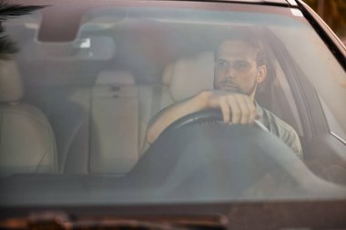 Genç bir adam bir arabanın içinde oturmuş, konsantrasyonunu bozmadan önüne bakıyor. İç kısım yavaşça bulanıklaşıyor ve sakin bir atmosfer yaratıyor.