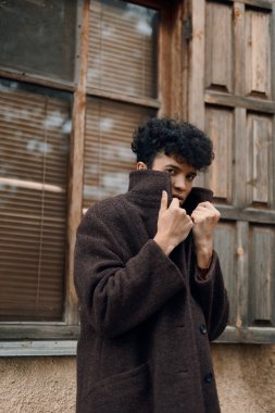 Kahverengi ceketli, elleri ceplerinde pencerenin kenarında kameraya bakan genç bir adamın moda fotoğrafı..