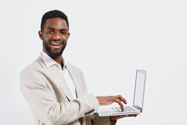 Koyu tenli neşeli bir adam, beyaz arka planlı bir stüdyoda dizüstü bilgisayarla çalışıyor. Açık renkli bir kazak ve beyaz gömlek giyer. Nişan ve kendine güven gösterisi yapar.