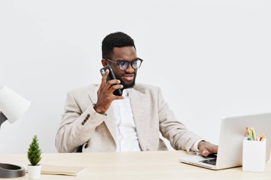 Gülümseyen Afro-Amerikan genç adam güncel ofis masasında çalışıyor cep telefonuyla iletişim kuruyor, dizüstü bilgisayar kullanıyor, minimal dekor, doğal ışık, kapalı alan...