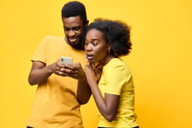 Neşeli genç bir çift, Afrika kökenli, uyumlu sarı tişörtler giyen, akıllı telefon sarı arka planına bakarken heyecanlı bir anı paylaşan neşeli bir atmosfer ve
